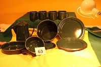 Full view of the enamel camp set showing coffee pot, deep pot, fry pan, bowls, mugs, and plates arranged on a yellow and green cloth background under warm lighting.
