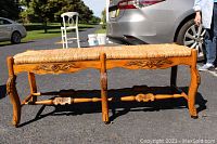 Front overview of the six leg bench showing the carved front apron with floral motifs and woven rush cord seat under daylight.