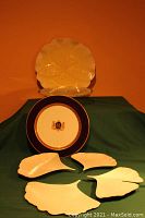 View of a white floral-shaped pie or cake plate, a navy-blue rimmed plate with gold emblem, and four white leaf-shaped plates arranged on a green cloth.