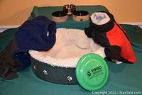 Overview of dog bed with ceramic and metal bowls on top plus two dog coats and a green frisbee