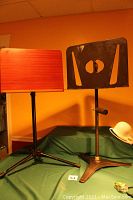Two music stands side by side: one with wooden surface and adjustable black tripod base; one with all metal design, black with cut-outs, and tripod base showing wear and rust.