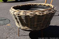 Large wicker planter on metal stand shown outdoors on pavement.