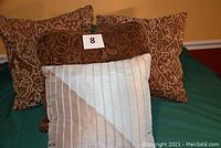 Four decorative throw pillows arranged on a bed showing both the front of the light aqua/beige pleated pillow and the brown patterned pillows behind it.