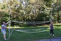 Volleyball net held horizontally by two people outdoors on grass with trees in background. Net is extended showing full length with poles on each end.