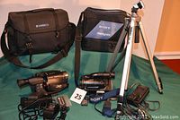 Full set photo showing Velbond tripod, JVC VHS camcorder with Ambico case, Sony Handycam with carrying case, and accessories arranged on table.