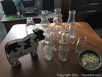 Twelve vintage clear glass milk bottles on wooden table with a black and white 5-piece wooden cow puzzle and a small crock-type bowl, plus paper bottle covers visible in bowl