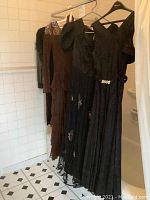 Four dresses hung on metal rack against tiled wall: black lace beaded-shoulder dress; brown lace tiered dress; black chiffon ruched dress with embroidery; black jacquard dress with rhinestone belt.