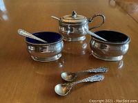 View of two sterling silver open salts with cobalt blue glass liners, mustard pot with lid and spoon, and five sterling silver salt spoons on wood surface.