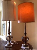 Pair of two tall lamps on table near window, one with white fabric shade and the other with smoked rust color shade.