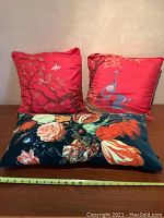 Front view of two red silk square pillows with cranes and floral patterns and one black velvet rectangular pillow with large colorful flowers, all on wooden surface with measuring tape
