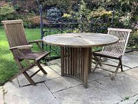 Two teak folding chairs and a round teak patio table placed on a stone patio with garden backdrop.