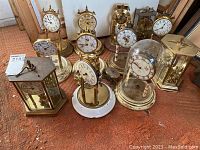 Group photo showing a variety of anniversary clocks, some under glass domes, brass framed, and several with floral designs on the clock faces.