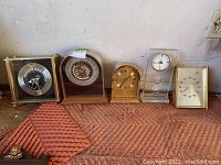 Five assorted clocks arranged side by side on a carpeted floor against a wall with electrical outlet visible.