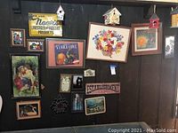 Wall with multiple framed wall art including vintage advertising prints, needlepoint floral basket, and small romantic images. Three painted birdhouses mounted high on the wall. Various signs including a yellow vintage poultry products metal sign.