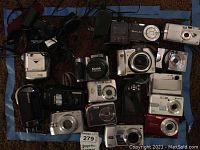 Photo showing multiple digital cameras of varying brands and colors arranged on a mat. Visible camera brands include Kodak, Samsung, Canon and more.