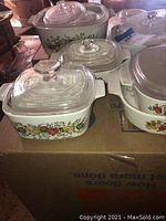 Multiple CorningWare ceramic baking dishes with vintage patterns including garden harvest and blue floral designs, each with glass lids, arranged on a cardboard box.
