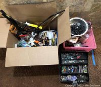 View from above showing an open cardboard box with various hardware items, a black multi-tier toolbox packed with small tools and fasteners, and a pink plastic container with additional hardware.