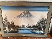 Framed oil painting showing a mountain, lake, and trees in muted blues and earth tones.