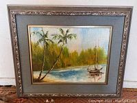 Framed oil painting with tropical lagoon scene, showing two palm trees and a sailboat, with some visible damage on canvas.