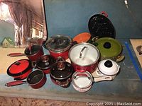 Assorted pots and pans displayed on a surface, mainly red cookware with visible lids and handles, some in black, white, and orange colors.
