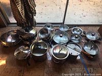 Assortment of various pots and pans laid out on the floor near a glass door, showing different sizes and lids.