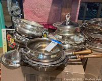 Photo showing various stacked silver plated bowls, serving dishes, and a small saucepan with wooden handle on cardboard box.