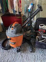 Ridgid orange/black shop vacuum with hose and power cord on carpet in front of mirror closet doors along with various small electronics in black bag.