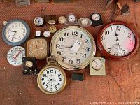 Overview photo showing a collection of various wall and desk clocks on a textured cloth surface. Clocks vary in size, style, and color.