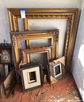 Collection of wooden frames of various sizes leaning against wall, some with decorative molding. One frame shows considerable damage with torn paper backing and wear. Nails visible on surface.