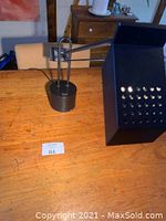 Photo showing the desk light from the side on a wooden table, highlighting the black weighted base and adjustable arm with rectangular perforated shade.