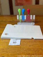 White rectangular ceramic cheese cutting board with two round corner holes and a side notch, placed on wooden surface with cheese knives stand in background