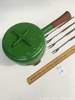 Bottom of kelly green fondue pot showing green enamel and wooden handle, four forks arranged next to a ruler.