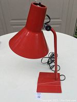 Front view of a mid-century red metal table lamp showing the conical lampshade, arm, and square base with electrical cord coiled behind.