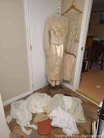 Full view of the Victorian wedding dress hanging with lace overlay and multiple undergarments displayed on floor.
