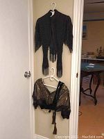 Two Victorian shoulder wraps hanging on white plastic hangers on a wall corner; one black beaded wrap with fringe and one brown-gold sheer wrap with velvet trim and tassel.