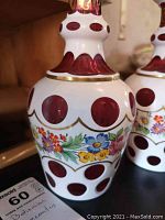 Close image of one decanter showing red cut to white glass with hand painted floral design and gold trim.