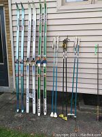 Full view of three sets of cross country skis and three sets of ski poles leaning against a house wall on a concrete surface.