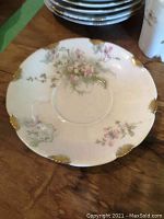 Close-up of demitasse saucer showing delicate floral pattern with pink flowers and gold trim at scalloped edges.