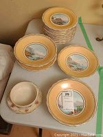 Stack of eighteen 9-inch plates and one smaller bowl on saucer shown with visible hand-painted scenic central design and golden-yellow rims.