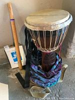 Photo showing djembe drum with natural skin head, shoulder straps, carry bag at base, rain stick standing next to drum, and music book on floor.