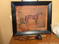 Full view of framed print of Nutwith horse with broken glass, showing wooden black frame and metal stirrups and horse hoof placed in front on table.