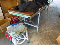 Wide side view showing the brown padded folding massage table with metal legs, black cover partially folded on top, plastic shower stool, and vintage muscle builder exerciser stacked