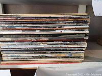 Stack of around 26 vintage vinyl record albums on a shelf, showing edge spines with artist names and wear.