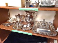 Full shelf view showing various silverplate pieces including gravy boat, pitcher, covered dish, tray, and flatware laid out on wood shelf.