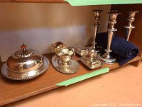 Silverplate covered dish, gravy boat with base tray, covered bowl with pumpkin-shaped handle lid, and four matching candlesticks arranged on wooden shelf.