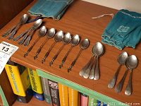Overview of all spoons on shelf including ornate Birks spoons, twisted-handle spoons, plain-handled spoons and two drawstring pouches