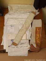 Top view of assorted cream and off-white linen tablecloths and napkins stacked with a rolled embroidered tablecloth on top; 'LINEN' labeled linen piece visible at right.