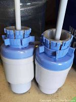 Two manual Dolphin water pumps, blue and white plastic, viewed from the side showing threads for bottle attachment and dispensing tube