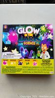 Front of the box showing the Glow In The Dark Science kit title with images of glowing experiments included and a cartoon scientist character.