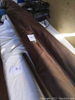 Brown patio umbrella rolled and wrapped in brown fabric cover lying on a white cloth surface. A close-up photo showing the umbrella cover and pole.
